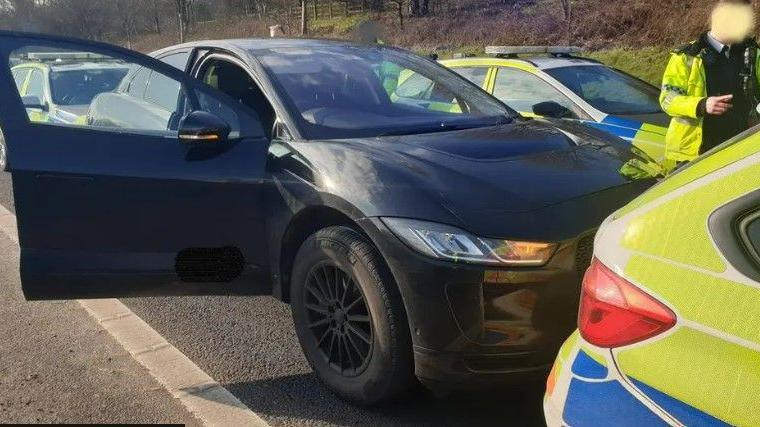 A black Jaguar I-Pace car parked on the motorway tightly boxed it at the front and rear by marked police patrol cars. An officer in a high visibility coat, with his face blurred, is standing on the passenger side of the vehicle. 