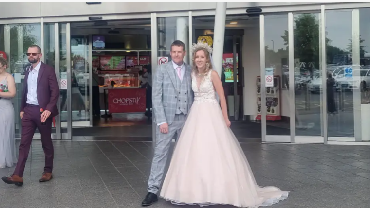 A groom and bride standing outside the service station. The bride is wearing a cream/ light pink wedding dress with a embroidery top. The groom is in a grey checked suit and a pink tie.  