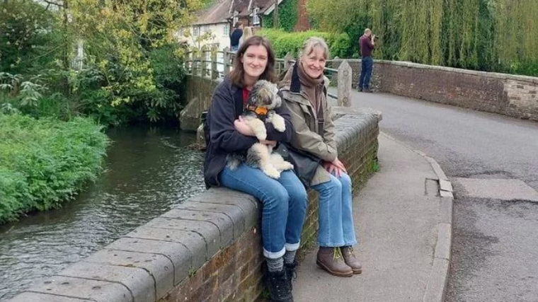 Two people, a mother and daughter, sat together on a wall next to a river, with a bridge and trees in the background. The daughter has fair skin and brown hair, and is cuddling a white and grey fluffy dog in her arms. Her mother is sat on her right, with fair skin and grey hair. They are both looking at the camera and smiling.