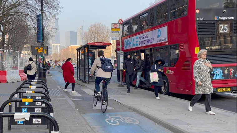 A pedestrian crosses a cycle lane to reach a floating bus stop, she is wearing a red coat and is stepping back slightly as a cyclist approaches in the cycle lane. On the right is a bus stop with a red London bus with passengers coming off.