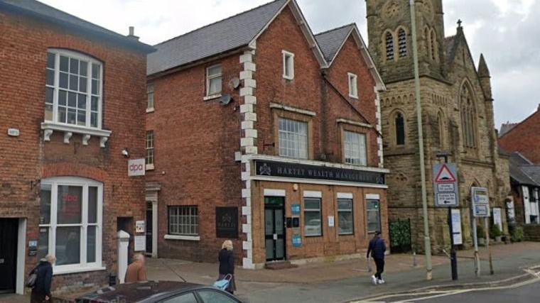 Two red brick buildings to the left of a stone church, with people walking on the street next to the buildings.