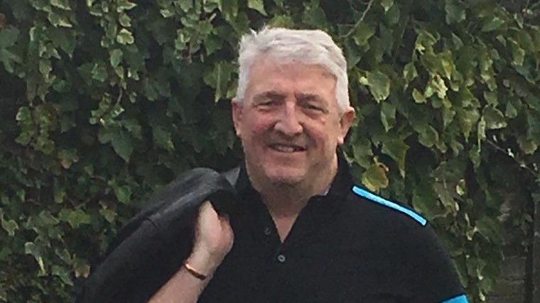 Paul stands outdoors for a photo. He has grey hair and is smiling at the camera