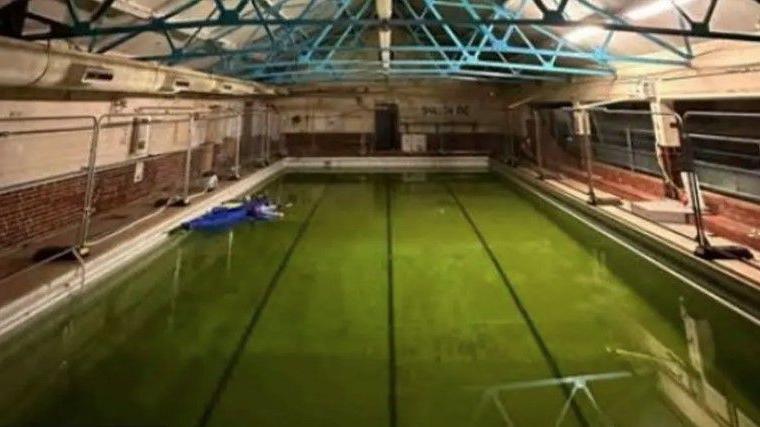 Indoor rectangular pool with four lanes, it's dimly lit, the water is dark green and metal fencing has been placed around its edge.