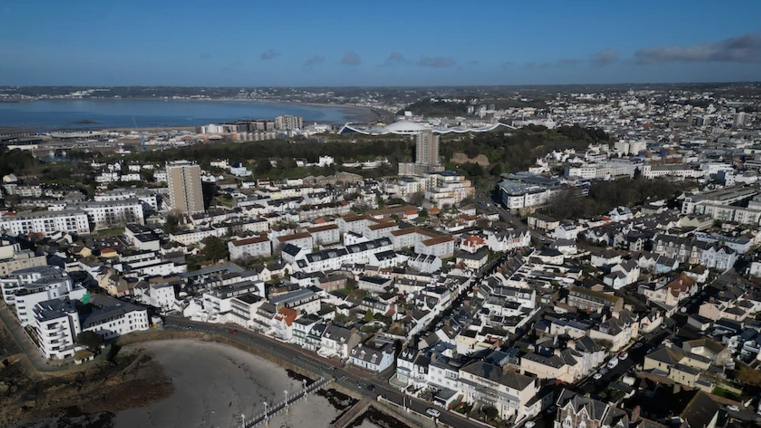 An aerial view of the south of Jersey - rows of houses and a beach in the foreground, sea can also be seen further afield. Blue skies.
