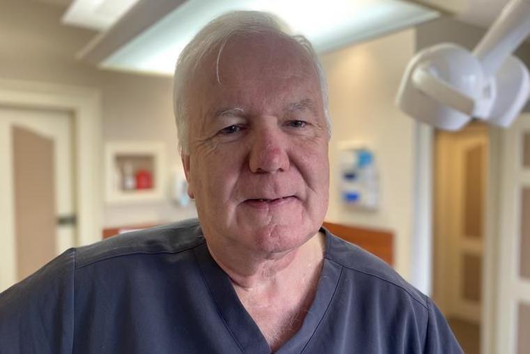 John Barry is an older man with white hair. He is in a dental surgery and is wearing a blue dentist uniform.