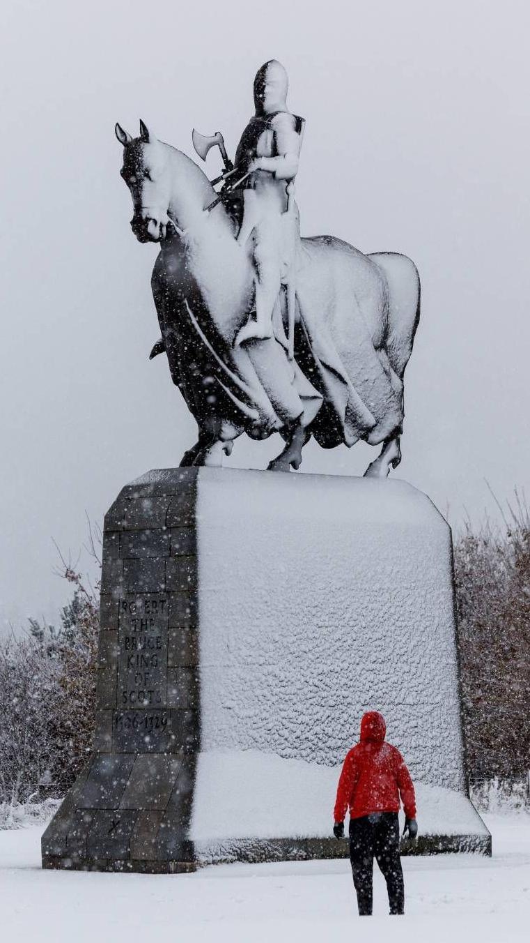 One side of statue of a man riding a horse on a large column covered in snow. A man wearing a red jacket, who looks very small in comparison, is looking up at the statue.