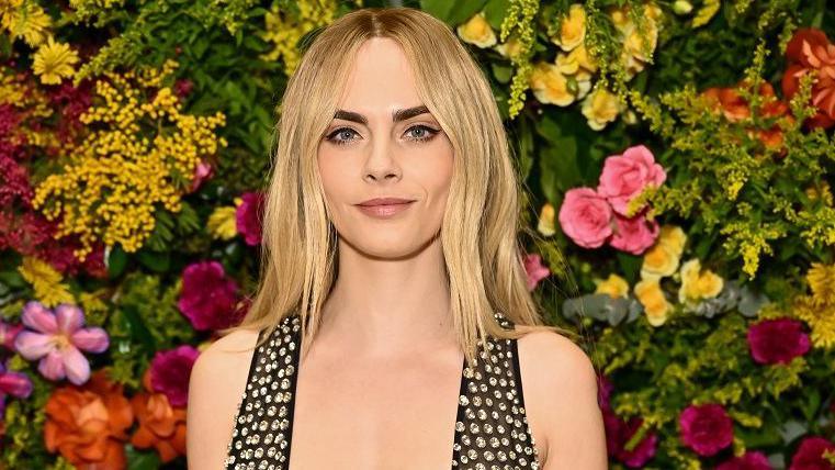 Cara Delevingne , with long blonde hair, stands in front of a floral display. The flowers are yellow, red, ornage and pink. She is smiling wryly, and wearing a black and silver dress