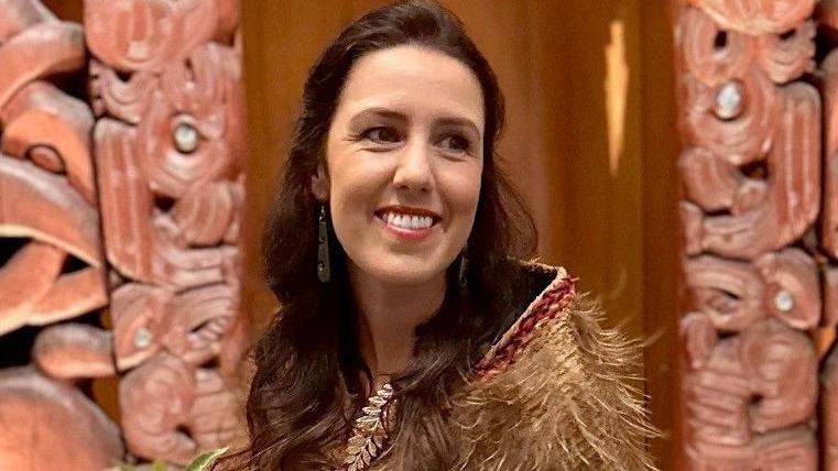 Laura wears a feathered cloak. She has long brown hair and is smiling. She is pictured in front of a decorative wall featuring Māori carvings.