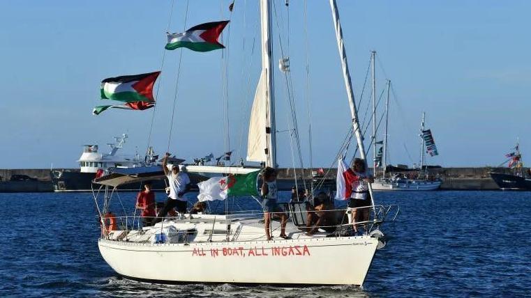 A boat that was part of the flotilla can be seen at sea with several Palestinian flags on it raised by the sails. The boat is small and white with "All In Boat, All InGaza" written in large red letters on the side.