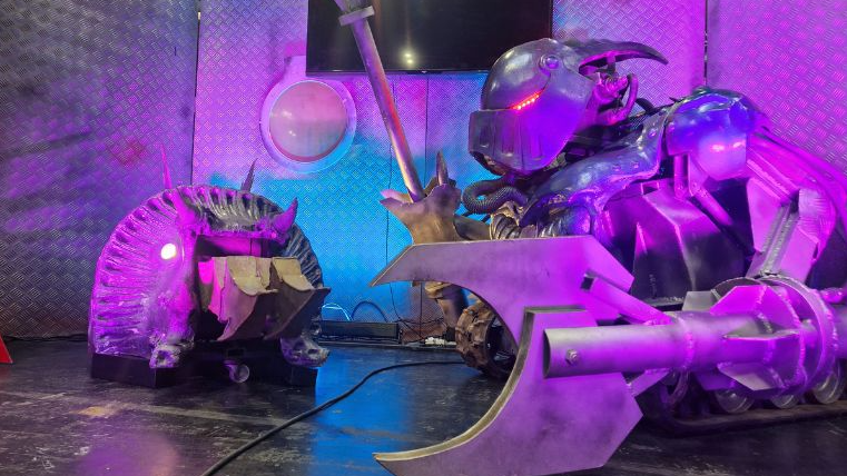 The two machines stand by one another in purple-tinged light. One looks like an armadillo with small bits of protruding metal from the front. A larger machine stands on the right which has a pincer on one side and hammer-type weapon on the other.