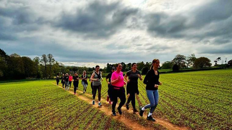 Woman run along a trail through farmland. There are trees in the background. Some of the runners are running side by side.