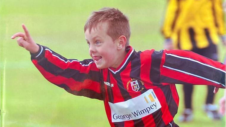 A young white boy with fair hair looks elated as he runs across a football pitch, smiling broadly with one hand in the air. He is wearing a black and red striped football kit and appears to be celebrating