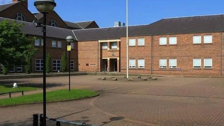 A pale-brick two-storey building with lots of windows and a bricked forecourt 