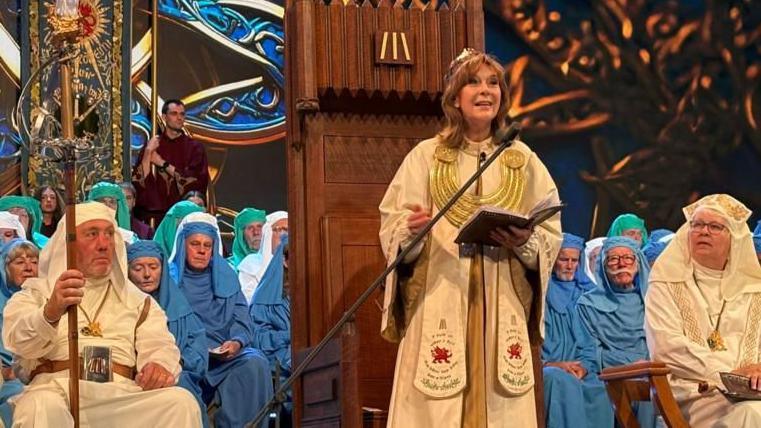 Archdruid Mererid Hopwood addresses new members of the Gorsedd in their official robes of green, white and blue. She is stood in front of a microphone with white robes on and holding a book. To the left and right of her are the new members, who are all seated.