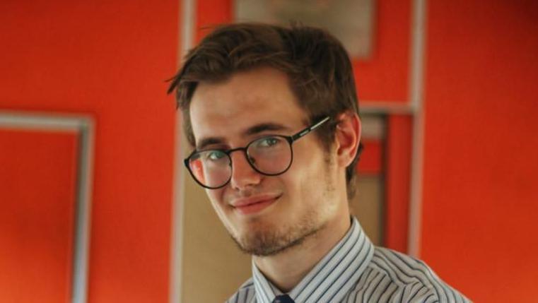 Brad Wright smiles at the camera while standing inside the Clacton Express' passenger carriage. He has short brown hair and is wearing black round glasses and a white shirt with dark stripes. 