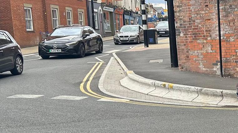 A view of the kerb on the corner with car having just gone past it. It is next to the large brick wall of the railway bridge. To the left, the street on one part of the junction with cars travelling down
