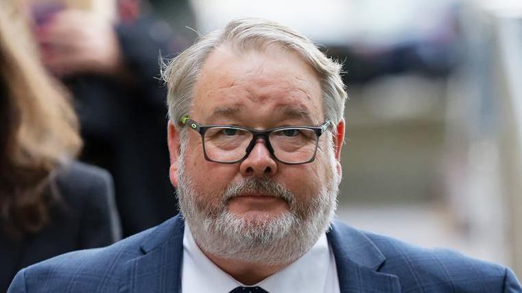 Close up of Christopher Mills outside court. He wears a blue suit, a white shirt and has black glasses. He has short grey hair and a grey beard. 