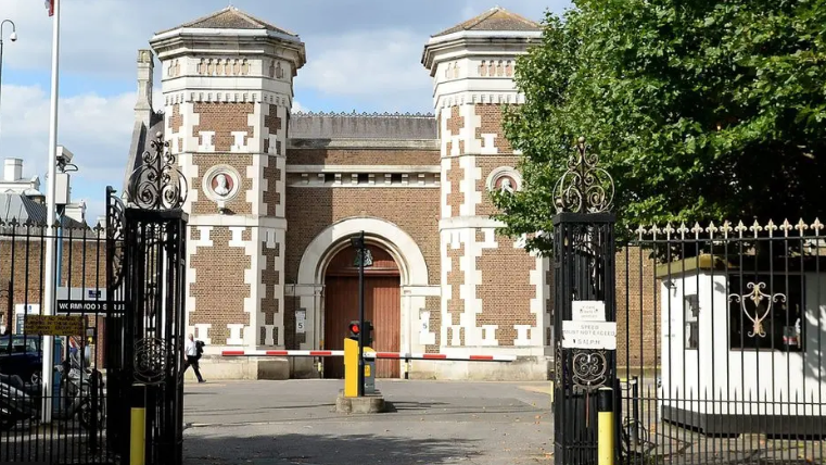 Entrance of Wormwood Scrubs prison, showing wrought iron gates, a car park barrier and a Victorian white and red brick prison with towers either side of an arched doorway 