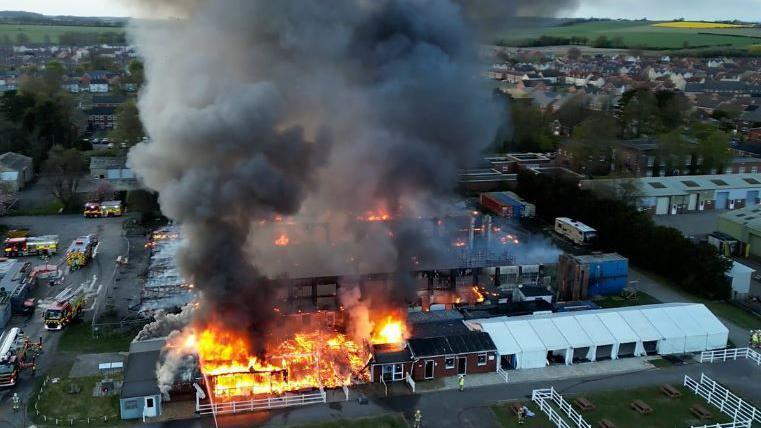 A black plume of smoke coming from a building on the airfield with a cordon and fire crews surrounding it