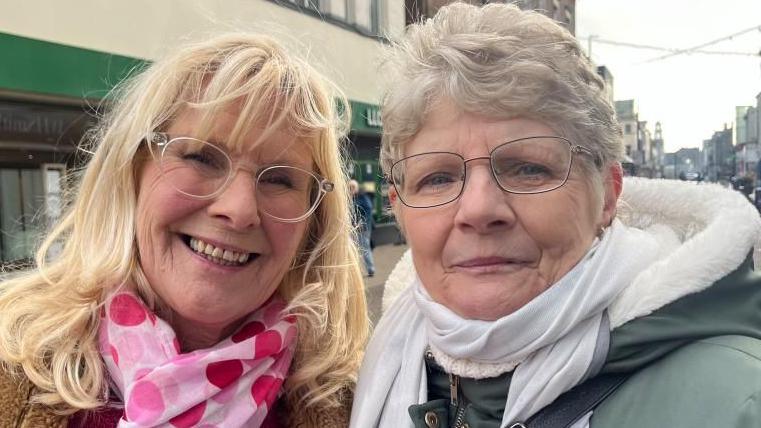 Amanda Chapman is on the left of the image, with long blonde hair. She is wearing a pink dotted scarf, a faux-fur lined jacket and a red jersey. Denise has greying hair, and wears glasses. She is wearing a white scarf, has a green jacket with white man-made fibre fur lining.