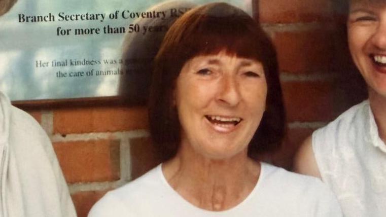 A dated picture of a woman smiling at the camera stood against a red brick wall. She is wearing a white t shirt and has short brown hair with a full fringe