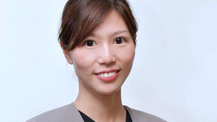 A professional woman smiling. She is wearing business like clothes and has dark brown hair tied back. 