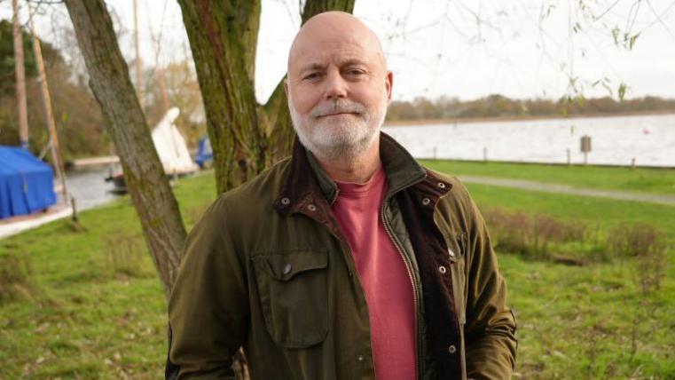 John Currie is wearing a green wax jacket and a light red T-shirt. He has a grey beard and is looking directly at the camera, standing on the banks of the Broads.