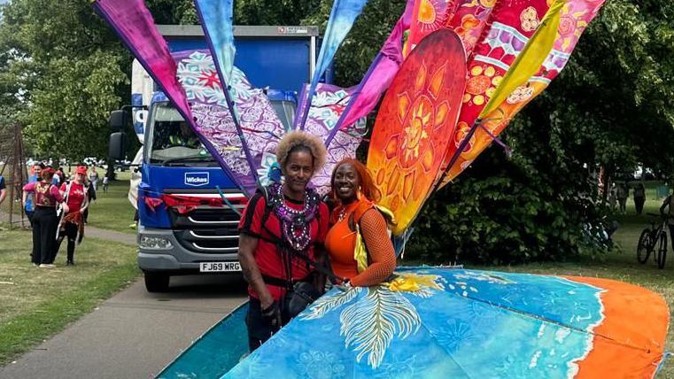 A man wearing a red T-shirt stands next to a woman in a colourful costume. It has a huge blue and orange skirt. Tied to her back are large banners that look like feathers and are red, orange and purple.