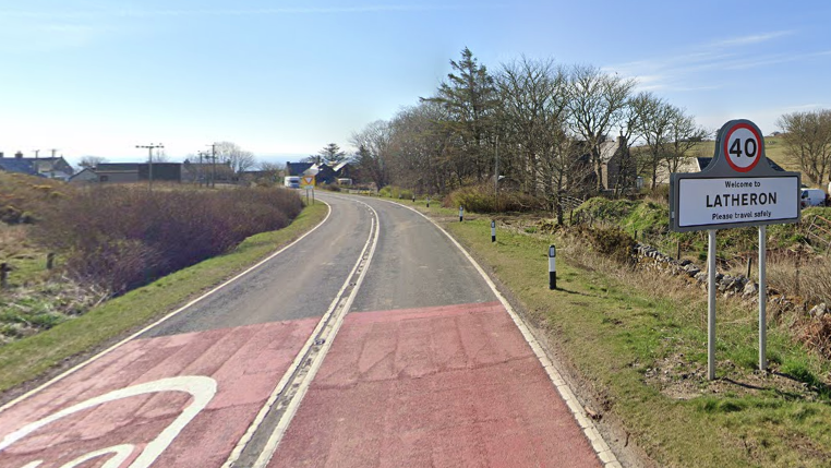A general view of the A9 road leading into the village of Latheron. A sign advertising the name of the village and encouraging motorists to drive safely is on a grass verge next o the road on the right.