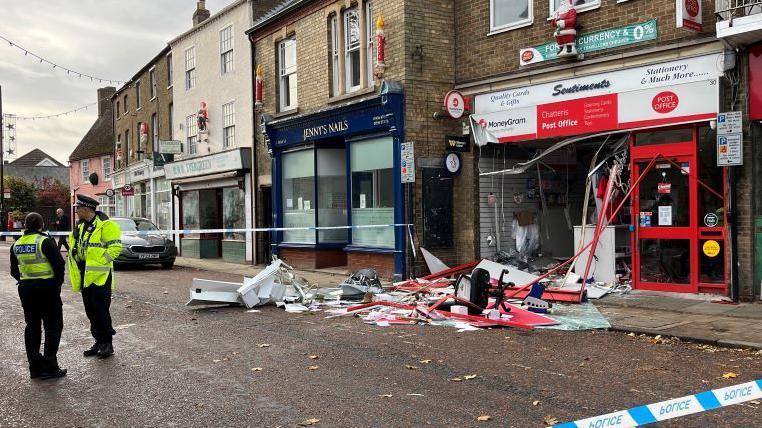 Damage to the front of Chatteris Post Office on High Street in the town. There is a police cordon in place with police tape around the front of the shop. Inside the cordon is two police officers. There is damage and debris from the shop splayed out onto the road.