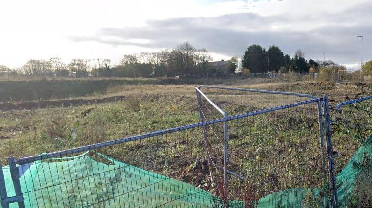 Google streetview of metal grid fence outside a section of green field with overgrown shrubs and tres in background.