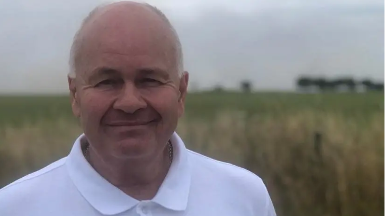 A head-and-shoulders shot of Neil Lockhart in a buttoned-up white polo shirt, stood in front of grassland and a field with trees silhouetted on the horizon under a grey sky.