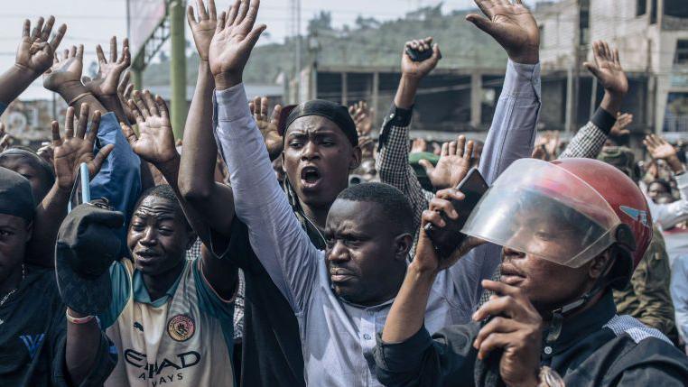 Goma residents, in a crowd, raise their hands as a soldier from the M23 group controls the access to the Stade de l'Unite (Unity Stadium).