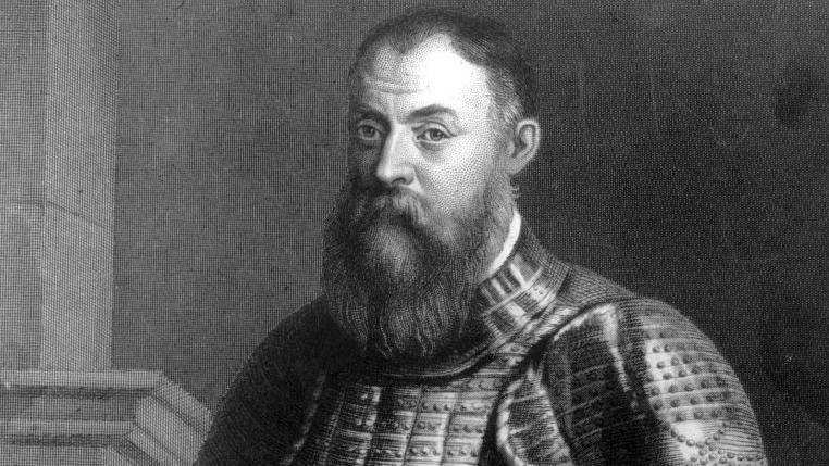 A black and white image of a man standing in military-type armour. 