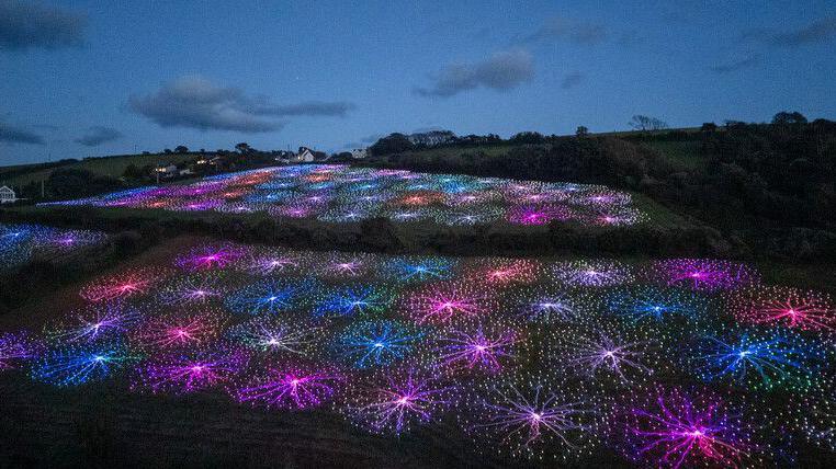 An aerial view of the Field of Light, the lights spiral out from various central points like a star. The work covers three fields, which are broken up with hedgerows. A dark sky can be seen in the background.