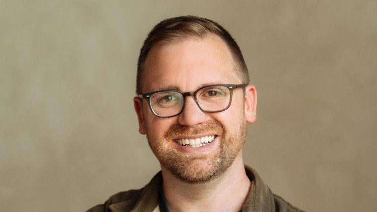 James Piper is pictured in a head and shoulders shot. He has brown hair and stubble and is wearing a khaki shirt and glasses. He is smiling widely.