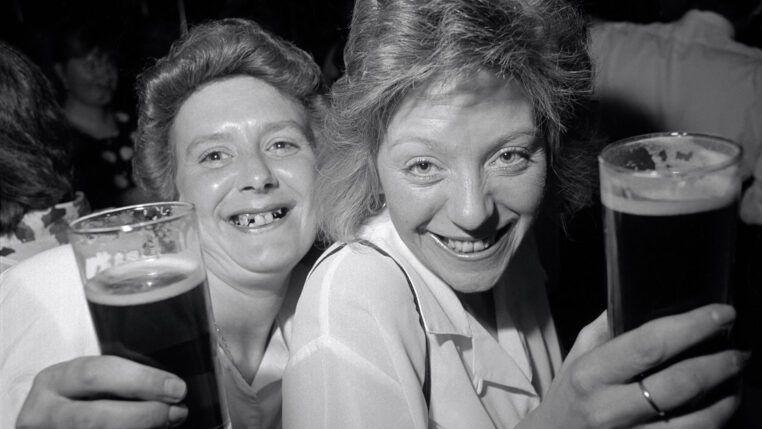 The black and white photo captures two people smiling and raising glasses of dark beer in a lively setting, looking directly at the camera.