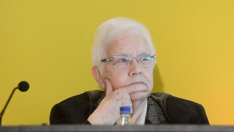 Baroness Blood is sitting in front of a mic. The backdrop behind her is yellow. There's a bottle of water on the table. Her hand is resting on her chin. She's wearing glasses.