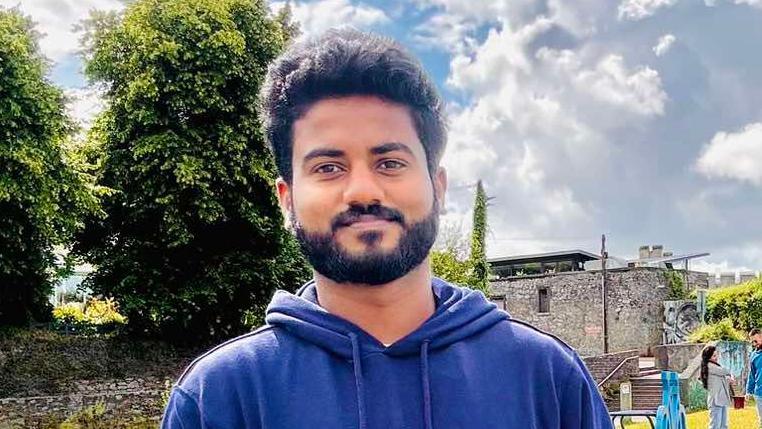 Man with a black beard wearing a blue navy hoodie standing in front of trees and a grey stone wall 