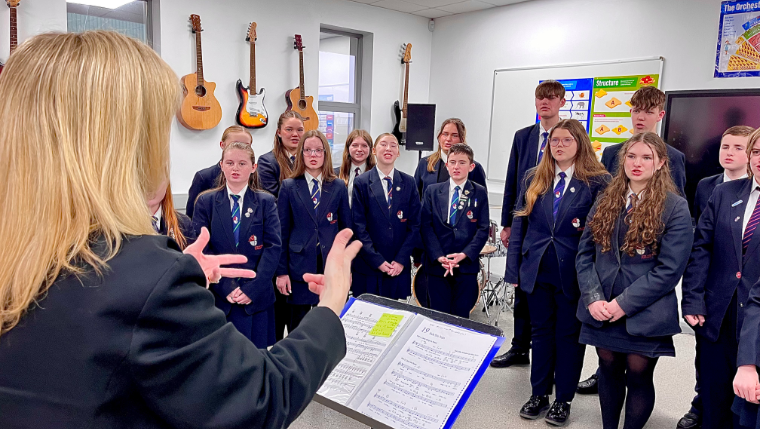 A woman leads a choir of teenage boys and girls dressed in navy school uniform in a music room, with guitars hanging on the walls