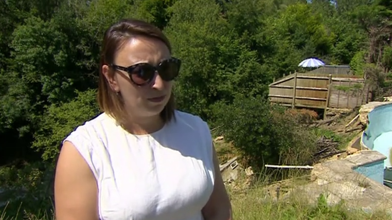 A white woman in a white dress with shoulder-length brown hair and brown sunglasses. She is stood in front of some trees. To the right is an empty swimming pool with a small amount of green water in it