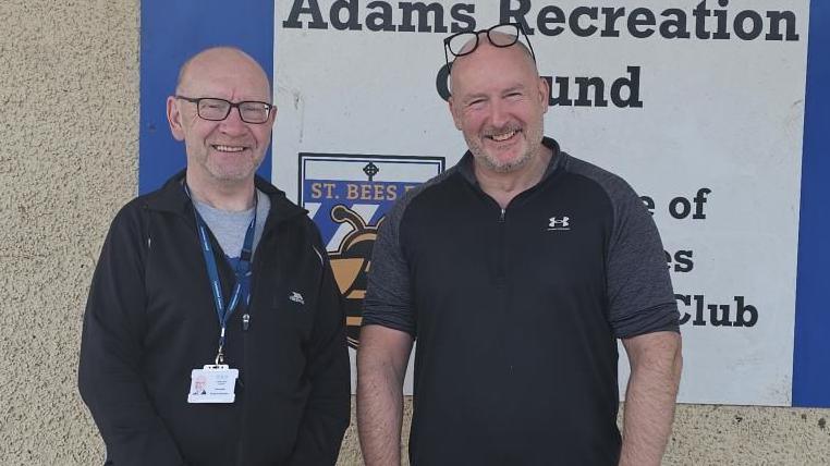 Councillor Graham Minshaw (Egremont North and St Bees) and Michael Rowell, Chair of Trustees for the Adams Recreation Ground, St Bees are smiling at the camera outside the recreation ground. Graham Minshaw is wearing a grey t-shirt, black sweatshirt, a blue lanyard and glasses. Mishael Rowell is wearing a black sweatshirt with grey sleeves. His glasses are on top of his head. There is a sign reading The Adams Recreation Ground on the wall behind them.