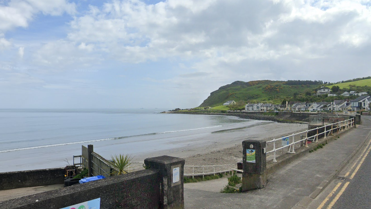 A beach seen from the road. In the foreground a low wall and then metal barrier. In the background houses along a headland with fields and trees behind them.