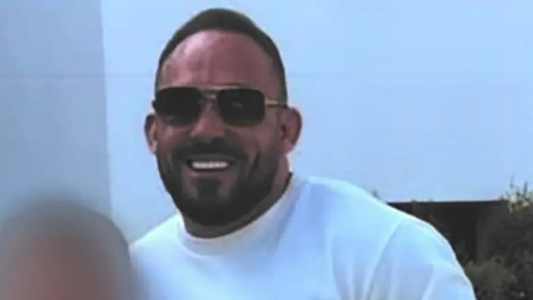 A photograph of Thomas Campbell, who has receding short brown hair, stubble, and is wearing sunglasses and a white t-shirt
