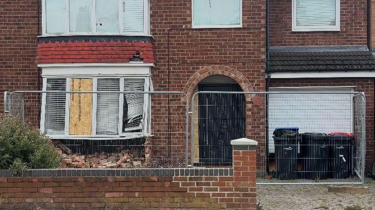 An alternate, close-up view of the damaged property. One of the smashed glass panes has been boarded up. A damaged set of blinds dangle from behind another pane.