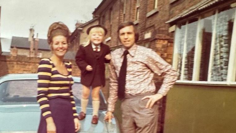 A family photo of a man and woman and a child, who is stood on a blue car, posing for a photo 