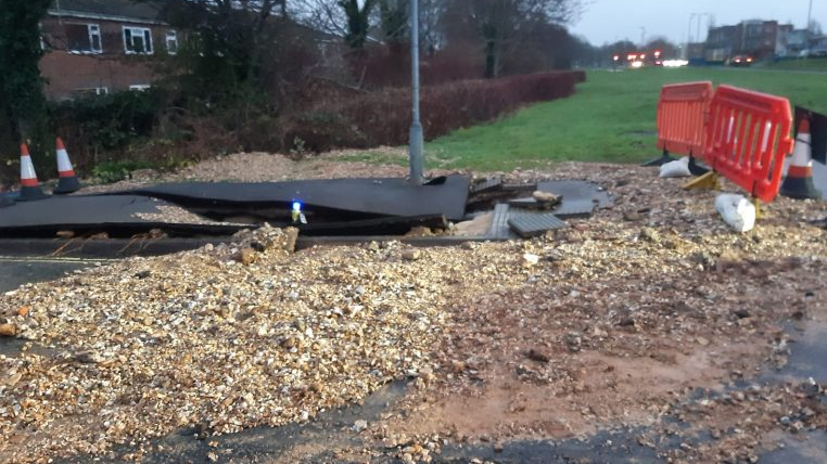 Damage left from a ruptured pipe, with waste and/or stone and silt washed up on the side of a path and clear damage to a path. A lamppost sits close by 