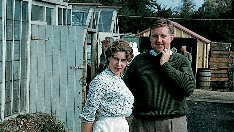 Hazel and Roger in 1960 standing outside the greenhouse, with a sign saying "Novelties and how to make the most of your dahlias". Hazel wears a pleated white knee-length skirt with a white and dotted black shirt tucked in. Her wavy hair is tied back. Roger wears grey slacks and a bottle green sweater. 
