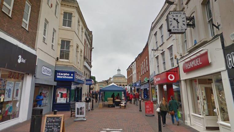 A pedestrian street with a line of shops on either side, including H Samuel and Carphone Warehouse. People are walking along the street.