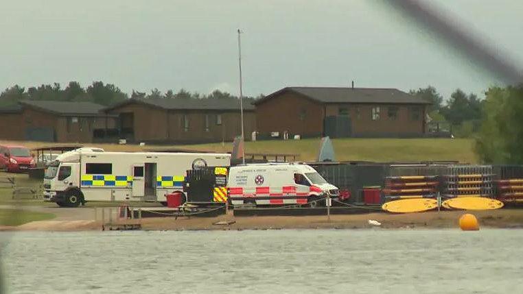 A lake in the foreground with a police mobile incident unit on the shore, a search and rescue team van next to it, two crates full of canoes also on the shore, and chalet buildings in the background.